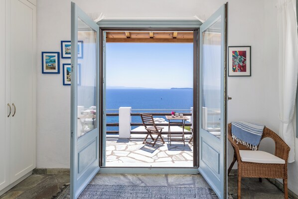 A room with a view of the sea, a wicker chair, a table, and a door leading to an outdoor area with a table and chairs.