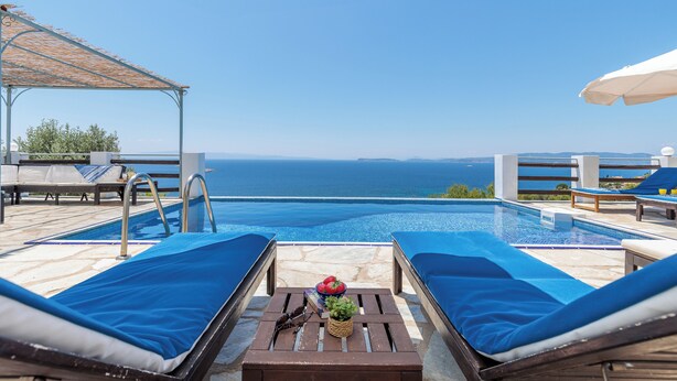 A poolside area with lounge chairs, a table with a plant, and a view of the sea. - Villa Kalyves Armenopetra - Beautiful  Pool - Amazing Sea Views -Walk to Beaches (Neo Klima)