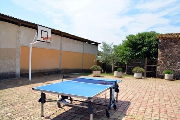 An outdoor ping pong table set up in a courtyard with a basketball hoop and potted plants.