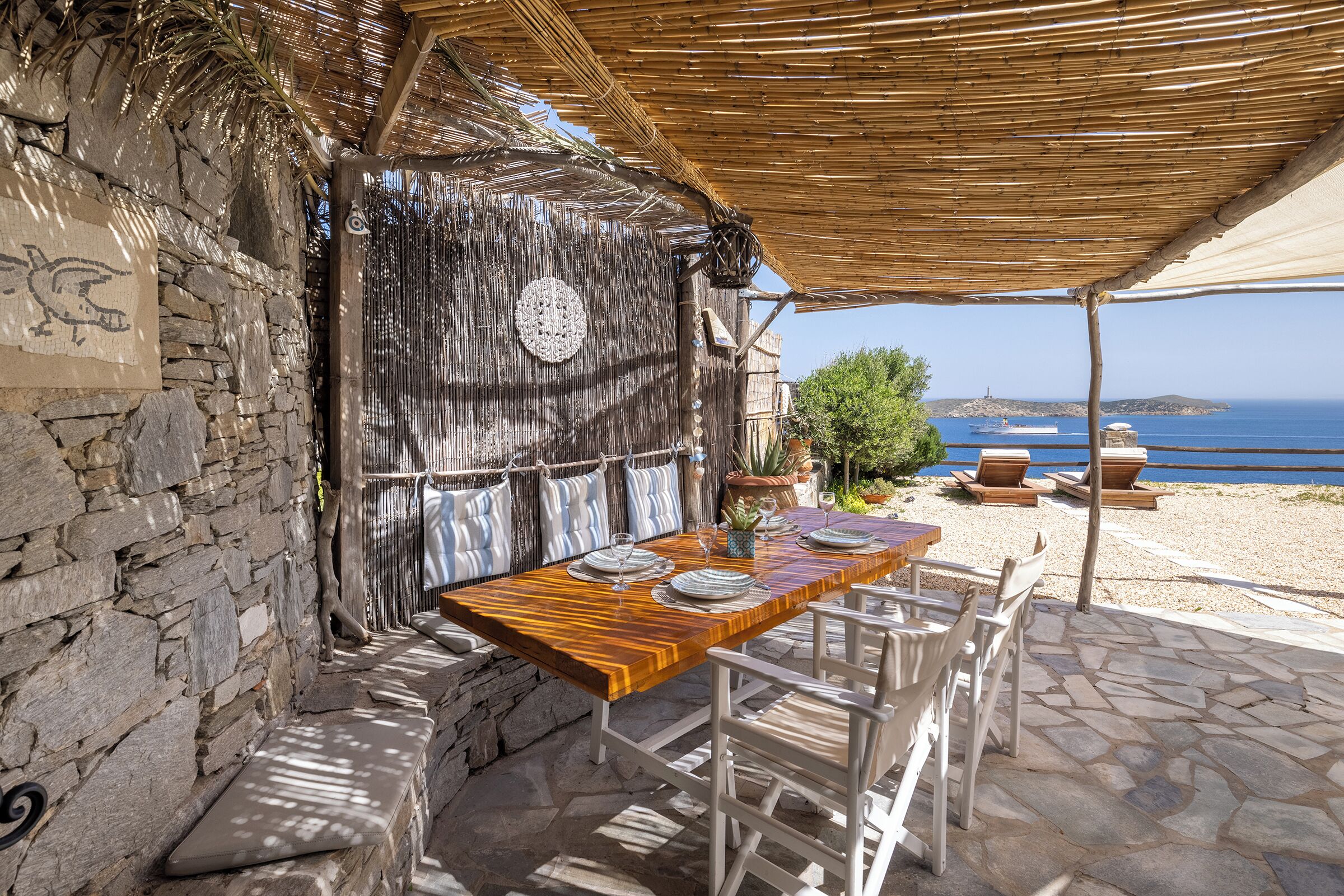 A stone-walled outdoor dining area with a wooden table and white chairs, overlooking a beach and the sea.