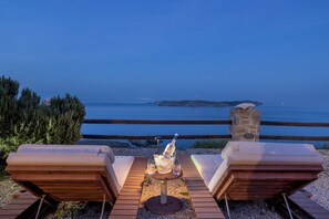 A wooden deck with two lounge chairs facing a body of water, a bottle of champagne, and two glasses on a small table. - Aegean View Seaside Apartment Syros (Syros)