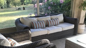 Ein Korbensofa mit Kissen, platziert in der Nähe eines Fensters mit Blick auf einen Garten.