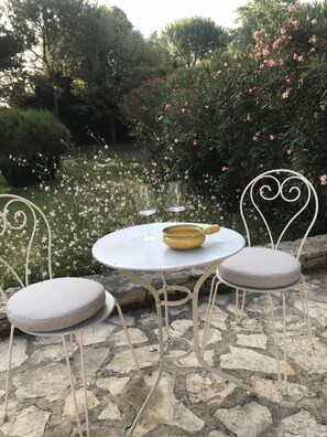 Une table ronde d’extérieur, surmontée d’un saladier jaune et accompagnée de deux verres à vin, est entourée de chaises en métal blanc dotées d’assises rembourrées. L’ensemble est installé sur une terrasse en pierre, avec un jardin en arrière-plan.