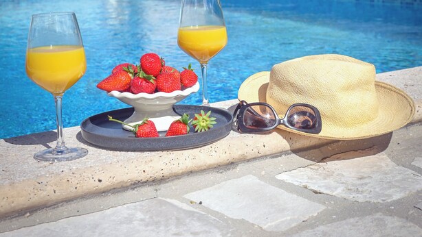 A poolside setting with a stone table, two glasses of orange juice, a bowl of strawberries, a straw hat, and sunglasses.