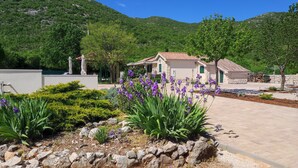A stone house with a tiled roof, surrounded by a landscaped garden with purple flowers and shrubs.