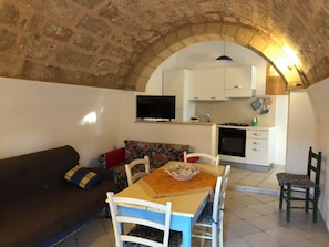 A cozy living area with a stone ceiling, a dining table with chairs, a sofa, and a kitchen with white cabinets and a flat-screen TV. - Villa CARLOTTA Magnificent Ancient CASEDDHU by the sea (Racale Torresuda)