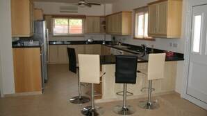 A modern kitchen with wooden cabinets, a black countertop, and two bar stools. - Luxury Three Bed House With Private Pool, Garden and BBQ (Agios Georgios)