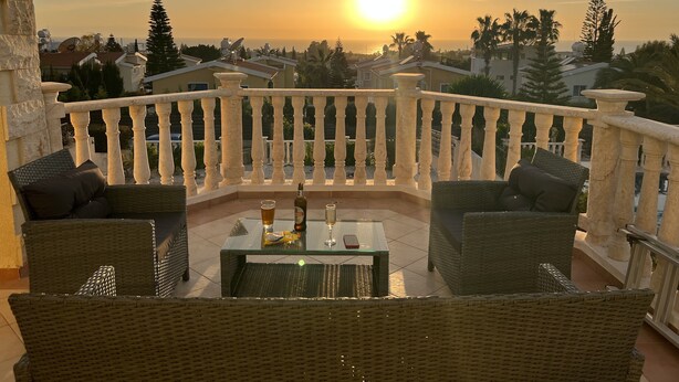 A balcony with wicker furniture, a glass table, and a bottle of wine. - Luxury Three Bed House With Private Pool, Garden and BBQ (Agios Georgios)