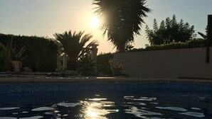 A swimming pool with sunlight reflecting on the water, surrounded by palm trees and a hedge. - Luxury Three Bed House With Private Pool, Garden and BBQ (Agios Georgios)