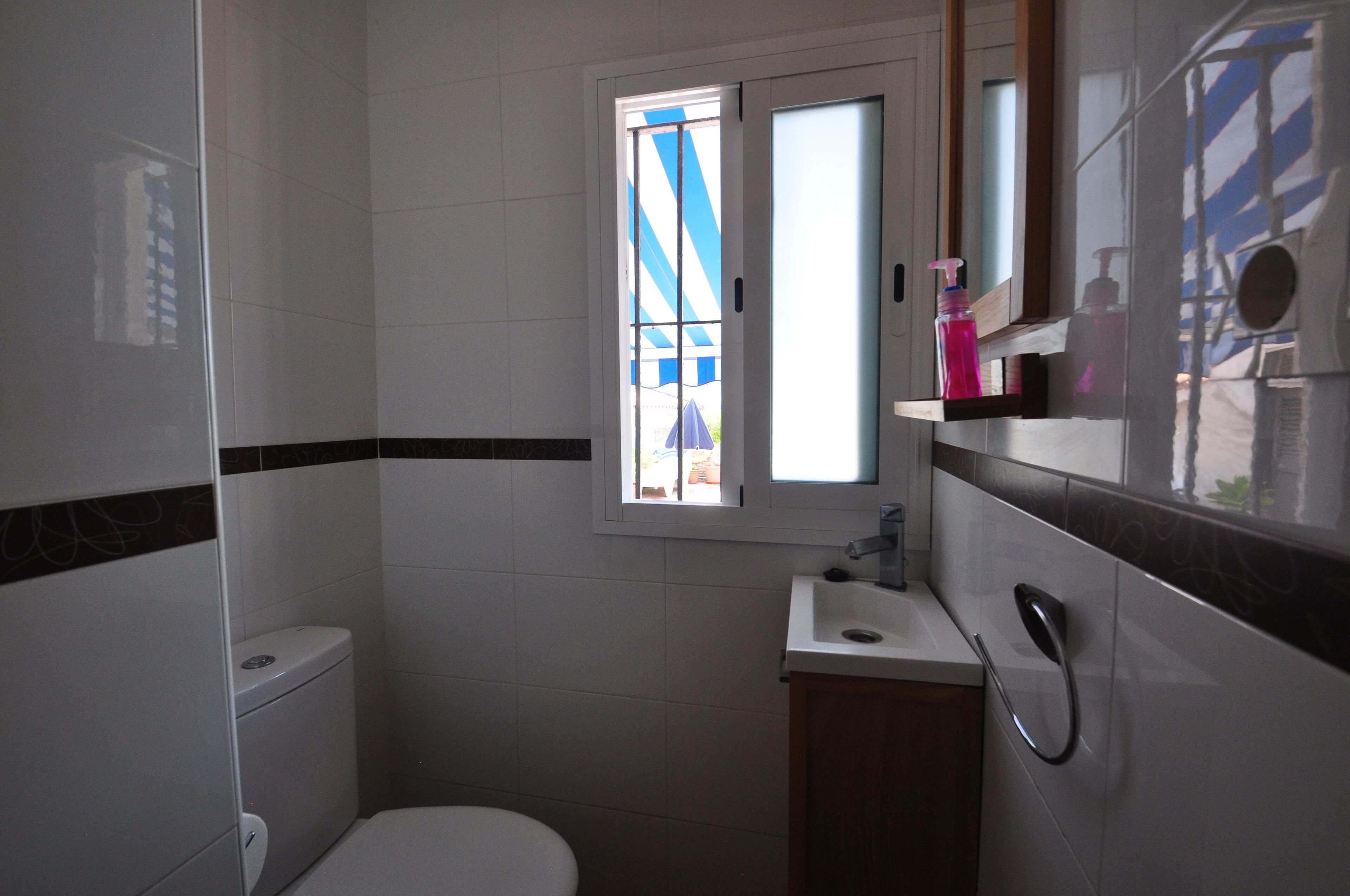 Un baño con inodoro, lavamanos, espejo y ventana con vistas a un toldo azul y blanco a rayas.