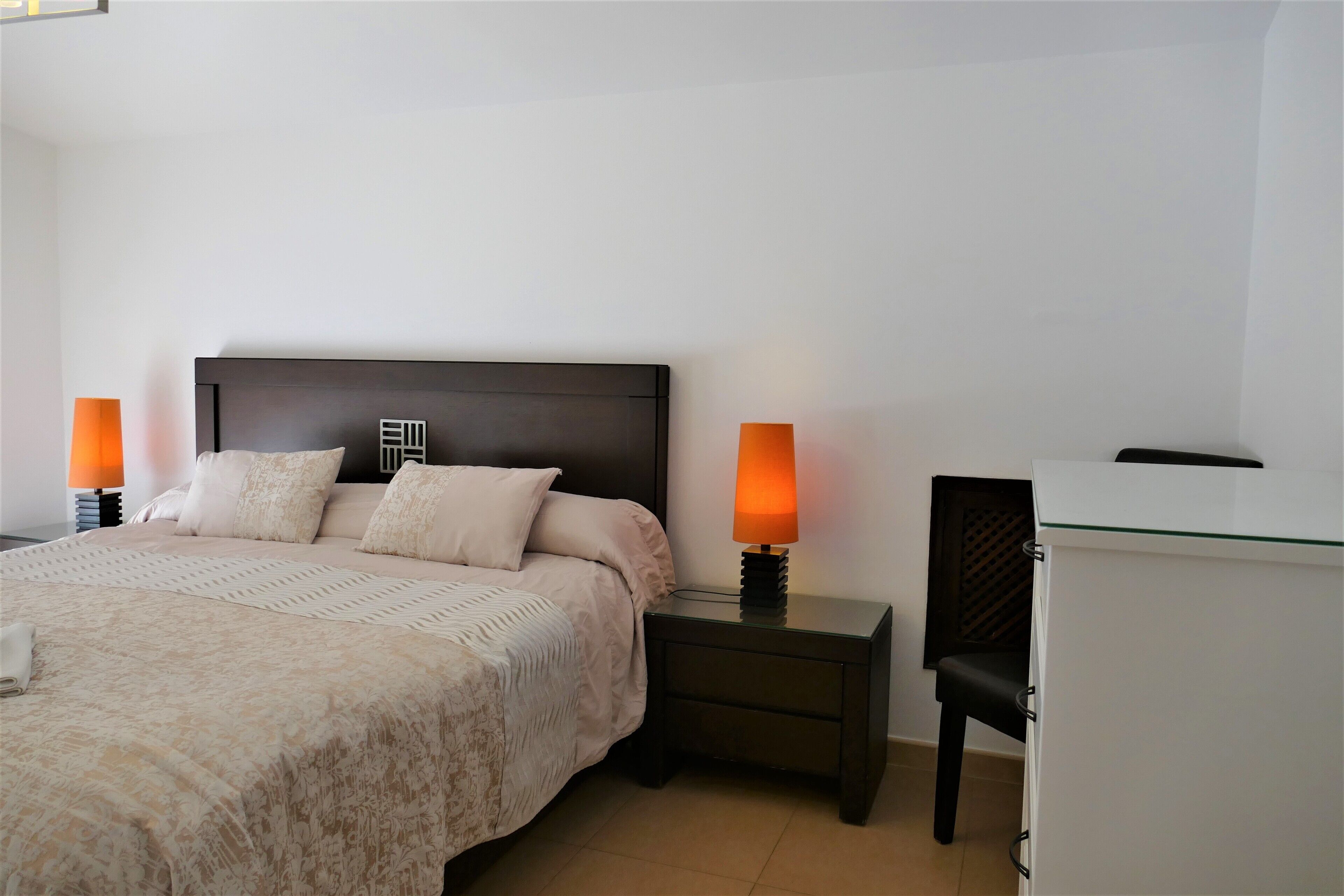 A neatly arranged bedroom with a large bed, two bedside lamps, a nightstand, and a small white cabinet.