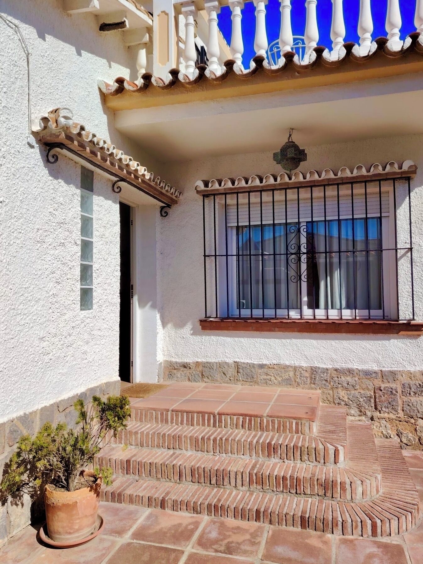A white building with a balcony, a window with metal bars, and a potted plant.