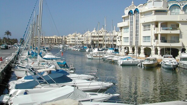 Un port de plaisance où les bateaux sont amarrés le long de la rive, entouré d’immeubles à plusieurs étages dotés de balcons.