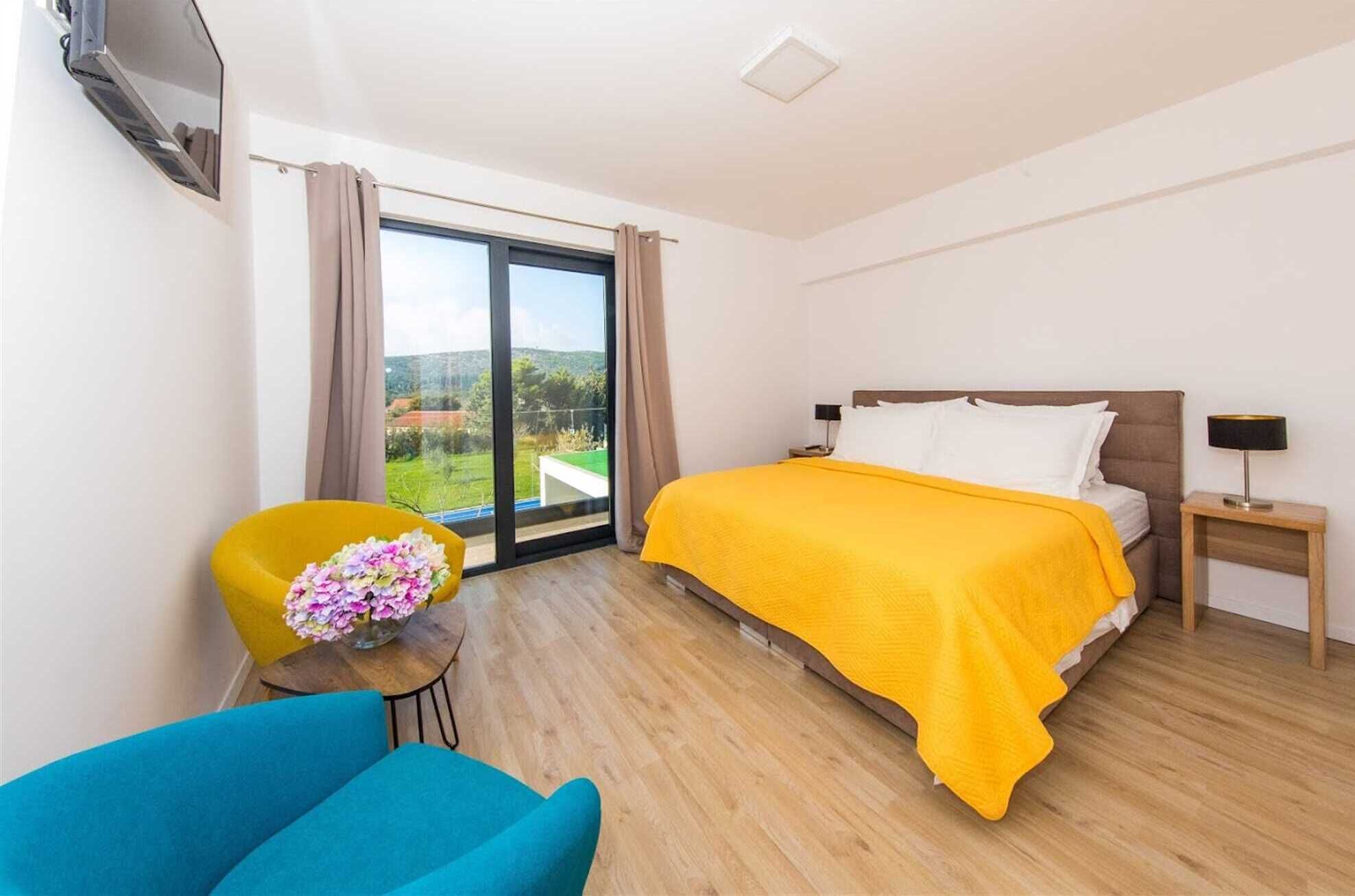 A hotel room with a bed, a chair, a small table with flowers, a nightstand, and a TV mounted on the wall.