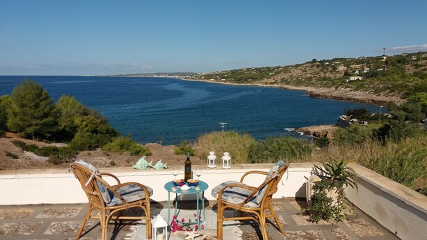 Eine Terrasse mit Korbstühlen und einem Tisch, mit Blick auf eine Küstenlandschaft, die einen klaren Meerblick und ferne Hügel bietet.