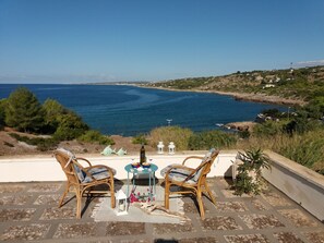 A terrace with wicker chairs and a table, overlooking a coastal landscape with a clear view of the sea and distant hills.