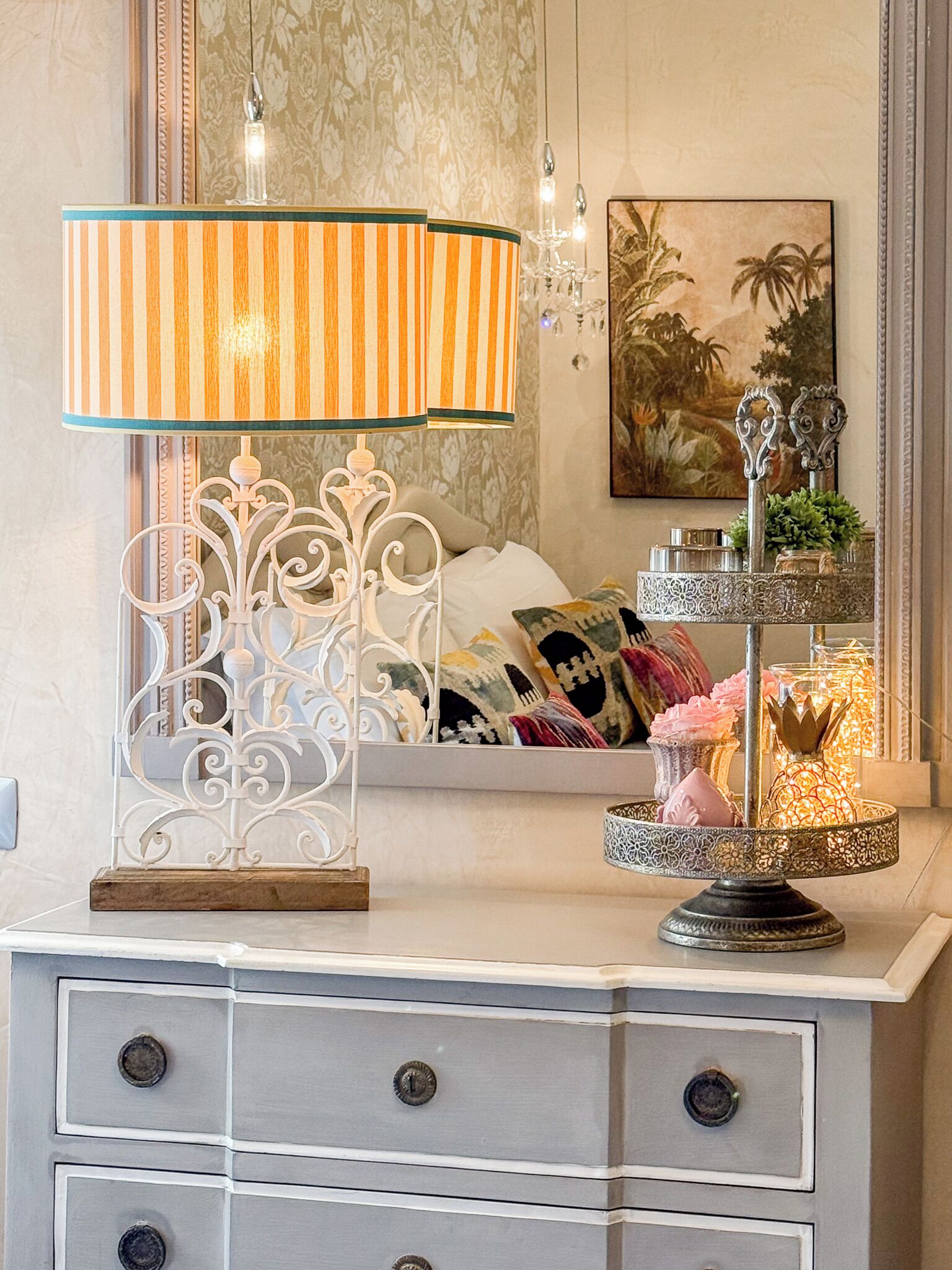 Une chambre à coucher comprenant un lit, une lampe de chevet, un miroir décoratif, une commode avec des tiroirs et une étagère murale agrémentée d’objets décoratifs.