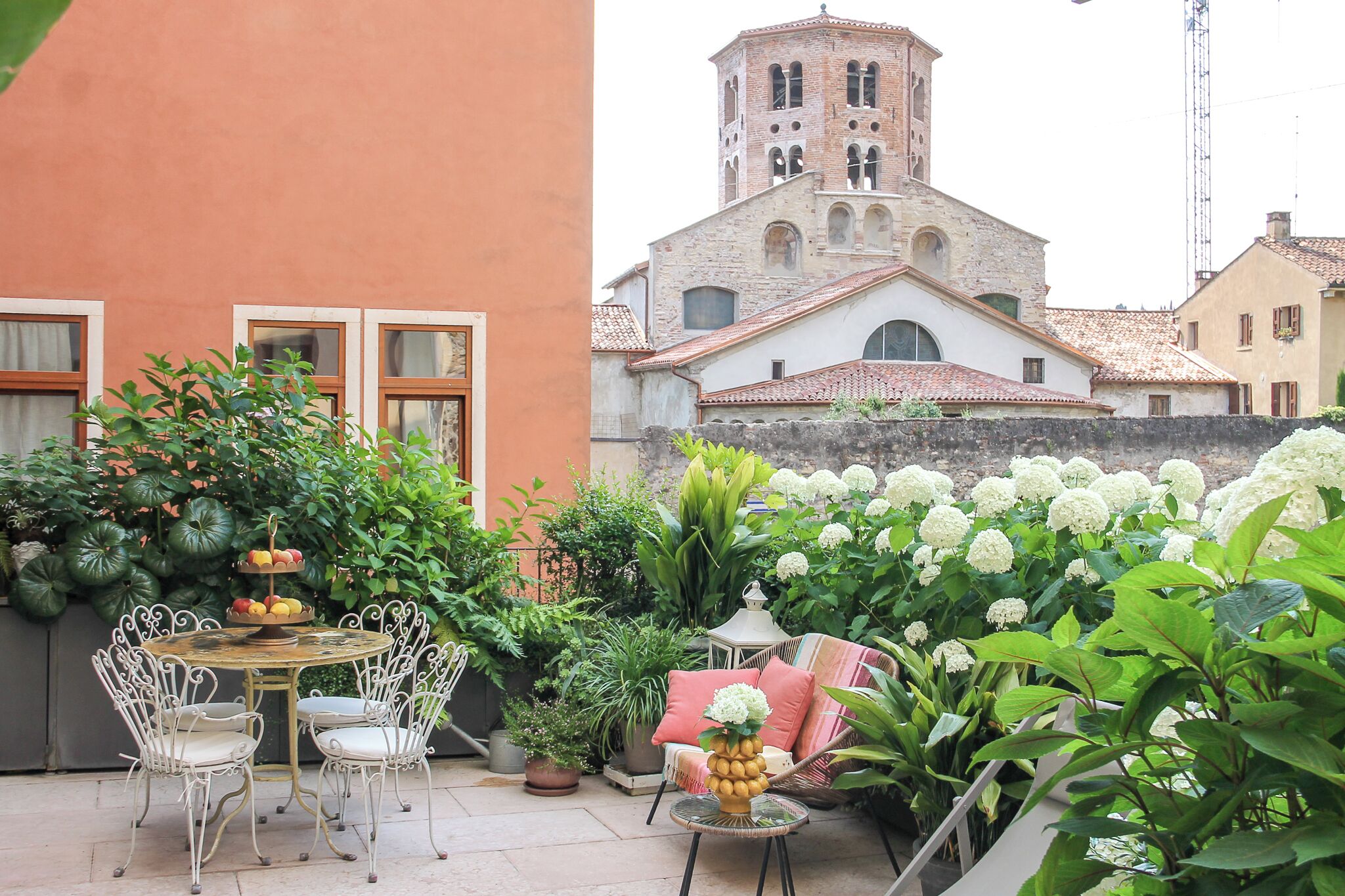 A courtyard with a round table and chairs, surrounded by potted plants and flowers.