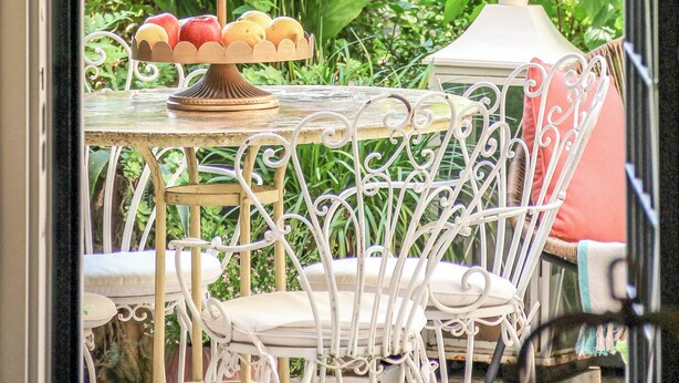 Una mesa de comedor con un frutero escalonado, sillas de metal blanco y vistas a vegetación y un edificio a través de una ventana.