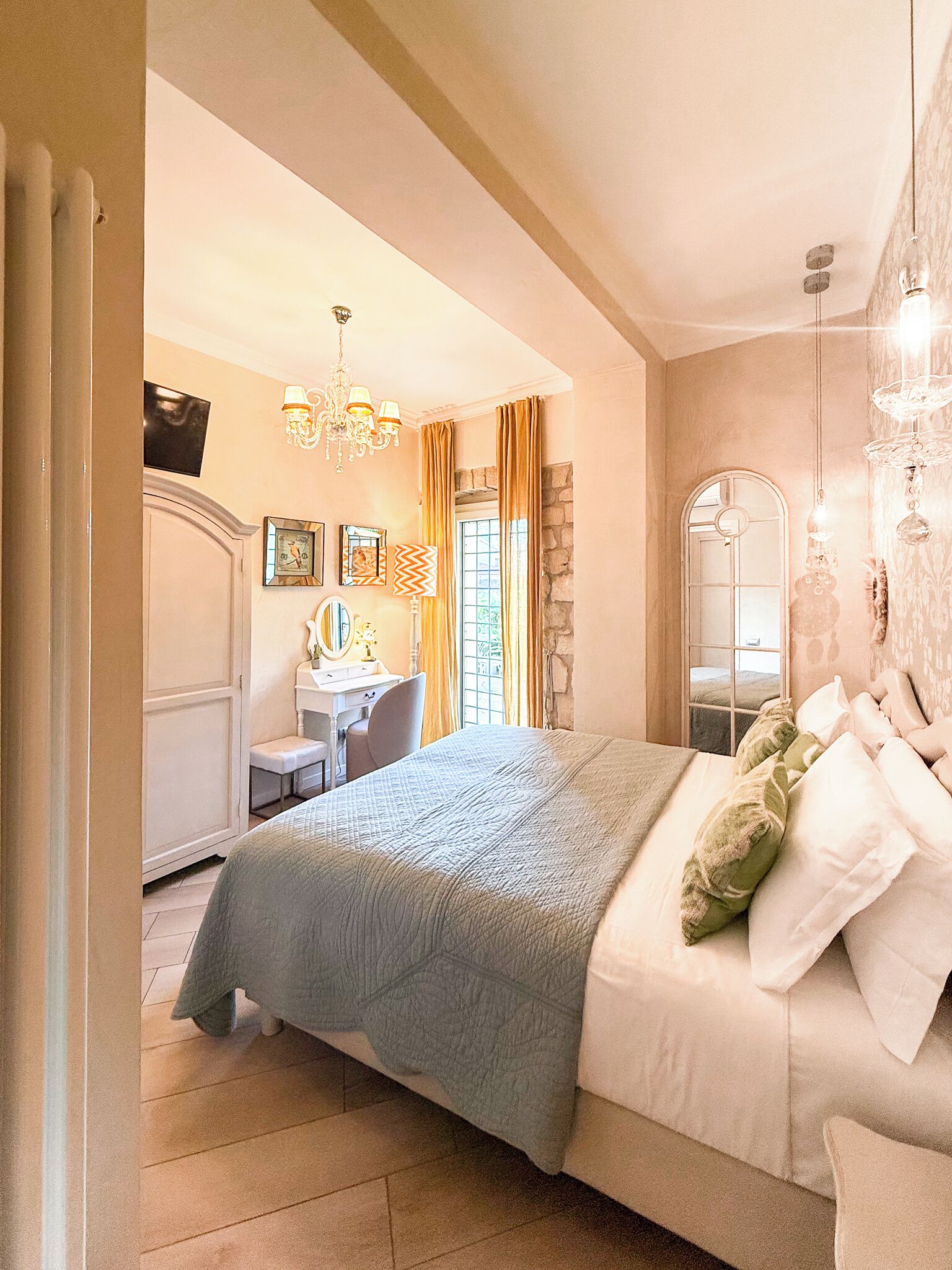 A bedroom with a bed, a desk, a chair, a mirror, and a chandelier.