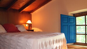 A bedroom with a bed, a nightstand with a lamp, a blue door, and a window with a view of greenery.