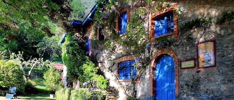 A stone building with blue doors and windows, surrounded by lush greenery.