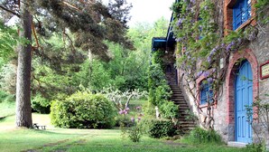 A rustic stone building with a blue door and window frames, surrounded by lush greenery and trees.