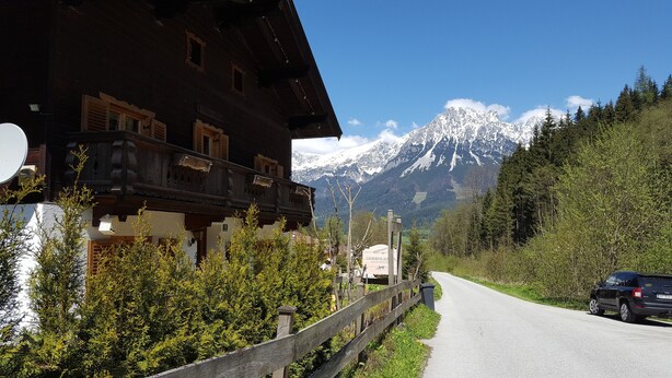 A traditional wooden building with a balcony, a road leading to a mountain, and a car parked on the side. - Ellmau: Quaint holiday home, 350 m to the mountain railway in the mountain doctor village (Ellmau)