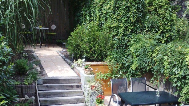 Eine Garten-Terrasse mit Tisch und Stühlen, umgeben von Pflanzen und einer Steintreppe.