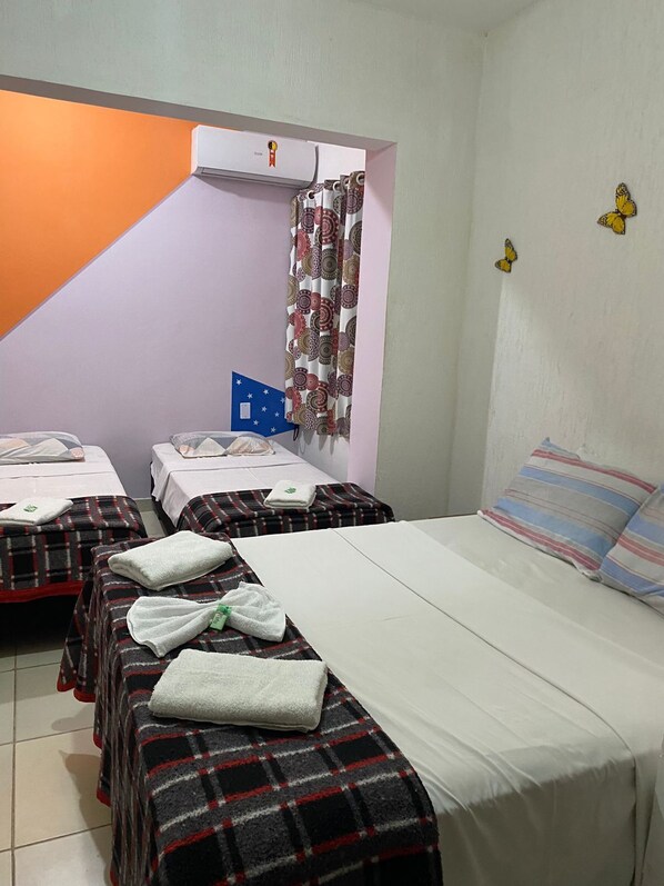 Quarto de hotel com duas camas, uma cama grande com almofada listrada e uma cama menor com almofada estampada. Há uma janela com cortina, ar condicionado fixado na parede e uma pequena bandeira azul na parede.