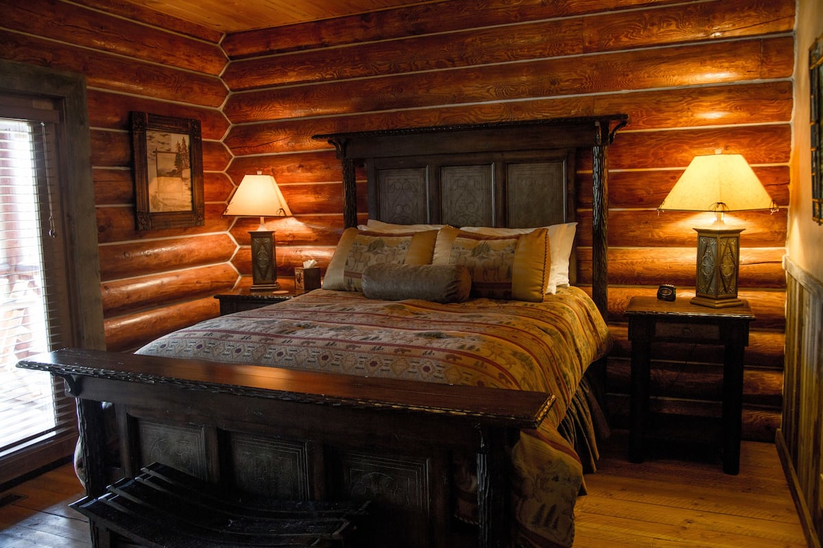 A log cabin-style bedroom with a large bed, two bedside tables with lamps, and a framed picture on the wall.