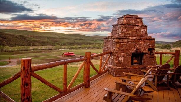 A rustic stone fireplace with a wooden deck and chairs, overlooking a scenic valley with a red-roofed building in the distance. - Ranches at Belt Creek (Belt)