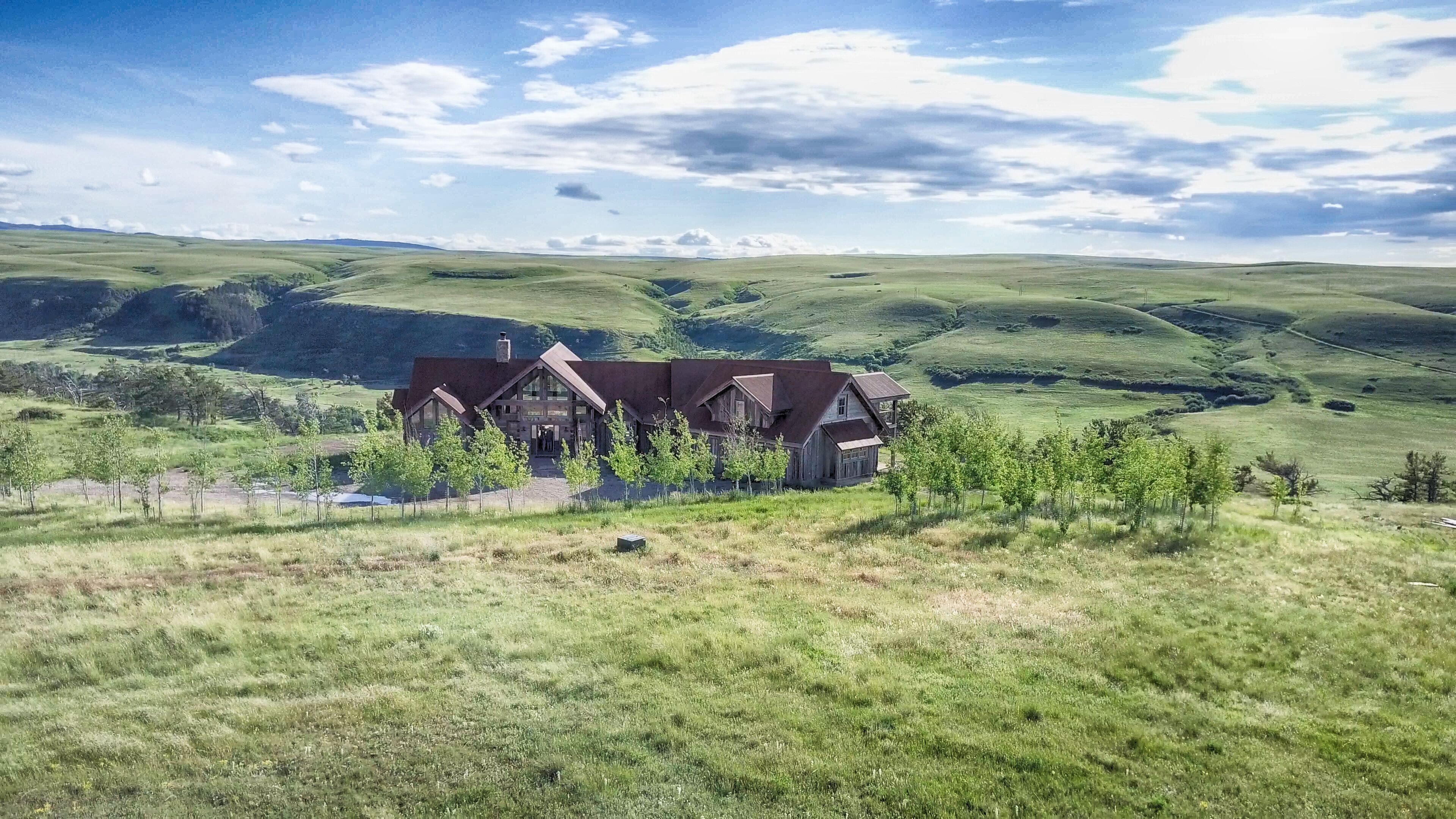 A large, rustic house with multiple sections, surrounded by rolling green hills and sparse trees.
