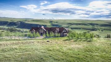 A large, rustic house with multiple sections, surrounded by rolling green hills and sparse trees.
