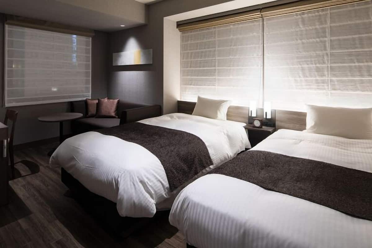 A hotel room with two beds, a small table, a sofa, and a window with blinds.