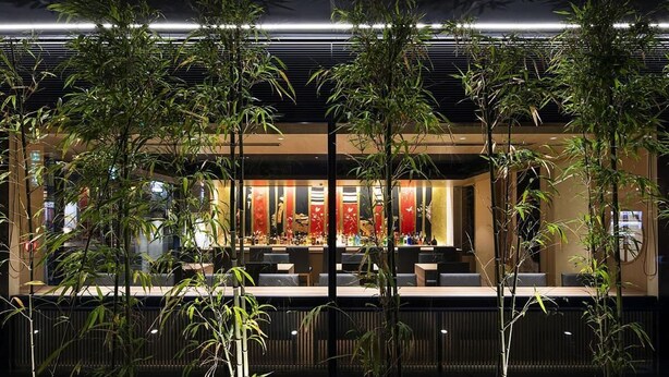 Un restaurante moderno con zona de bar, plantas de bambú e iluminación interior cálida.