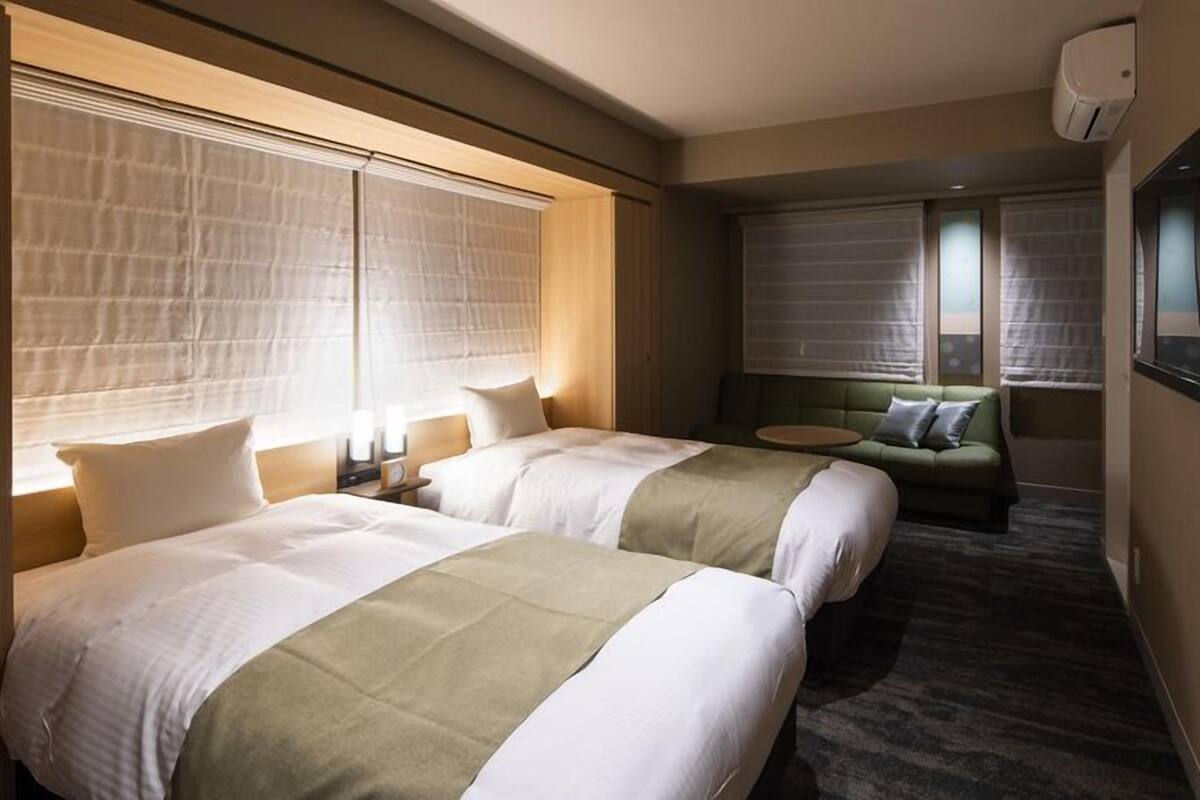 A hotel room with two beds, a sofa, a TV, and a window with blinds.