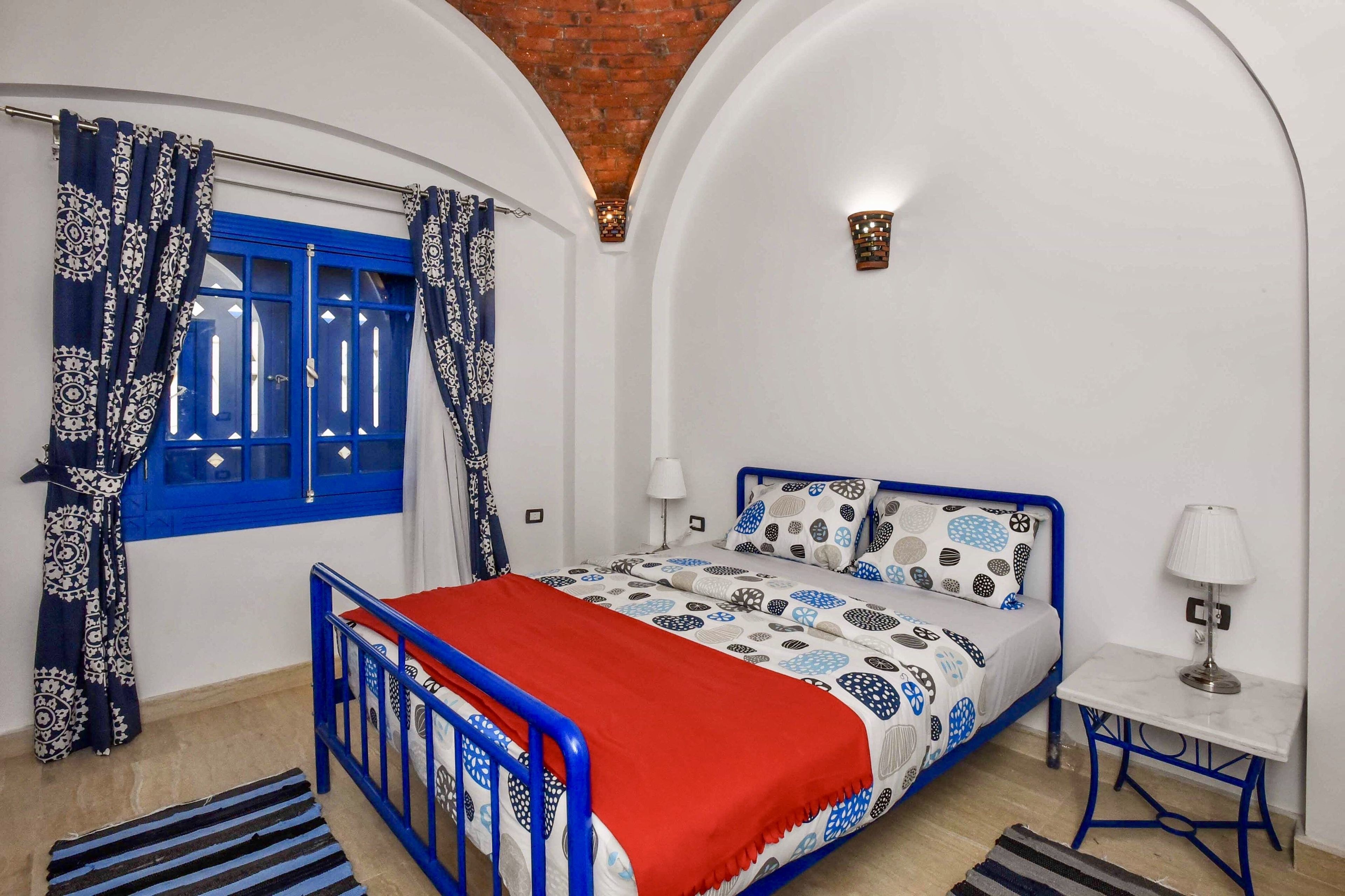 Une chambre à coucher avec un lit, une table de chevet, une lampe, une fenêtre avec des rideaux bleus et un plafond en briques.