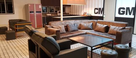A modern living room with a sectional sofa, a coffee table, and ottomans. There is a kitchen area in the background with a refrigerator and cabinets.