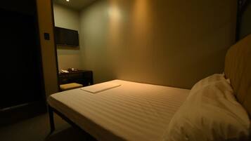 A small, dimly lit room with a single bed, a nightstand, a television, and a door.