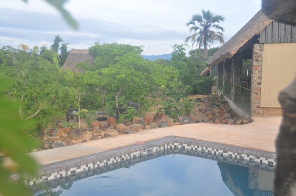 A pool area with a tiled edge, surrounded by rocks and greenery, and a building with a thatched roof. - Rafiki Safari Camp (Chikombe)