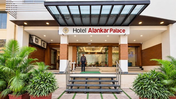 The entrance of Hotel Alankar Palace with a modern design, featuring a glass canopy, a reception desk, and a staircase leading to the entrance.