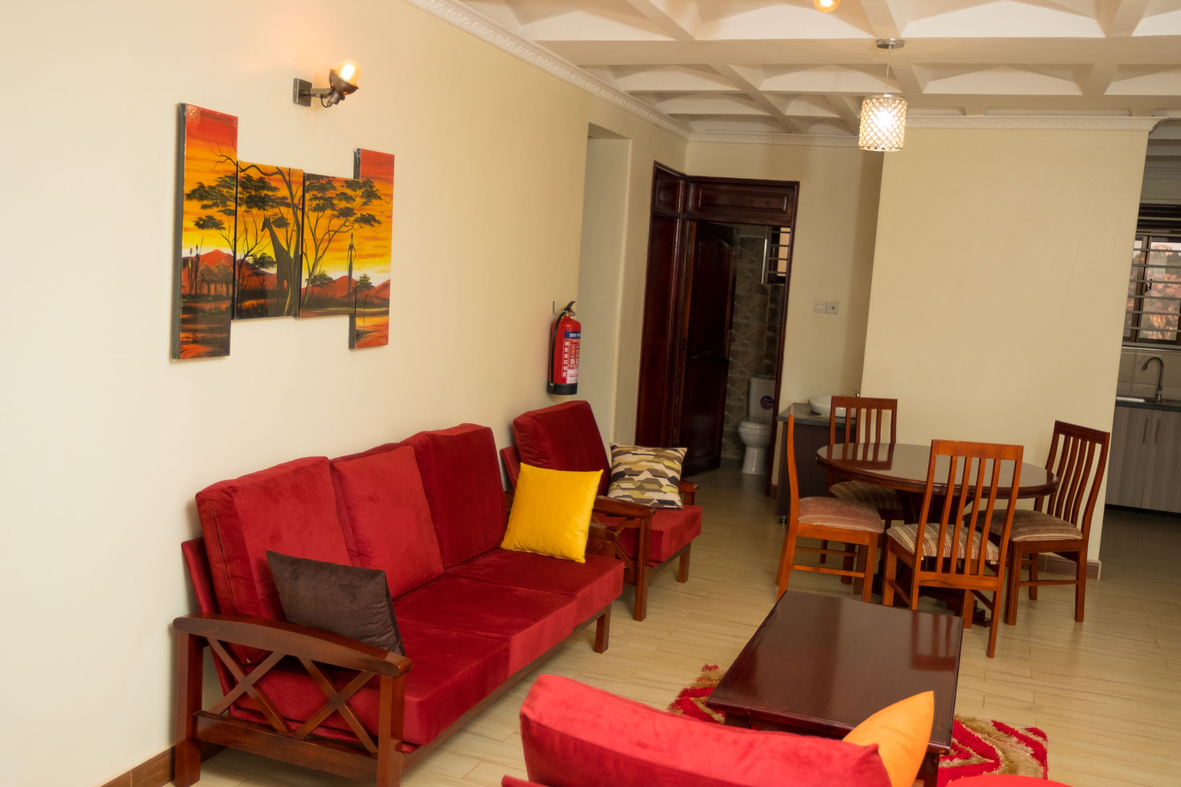A living room with a red sofa, wooden chairs, a dining table, and a kitchen in the background.