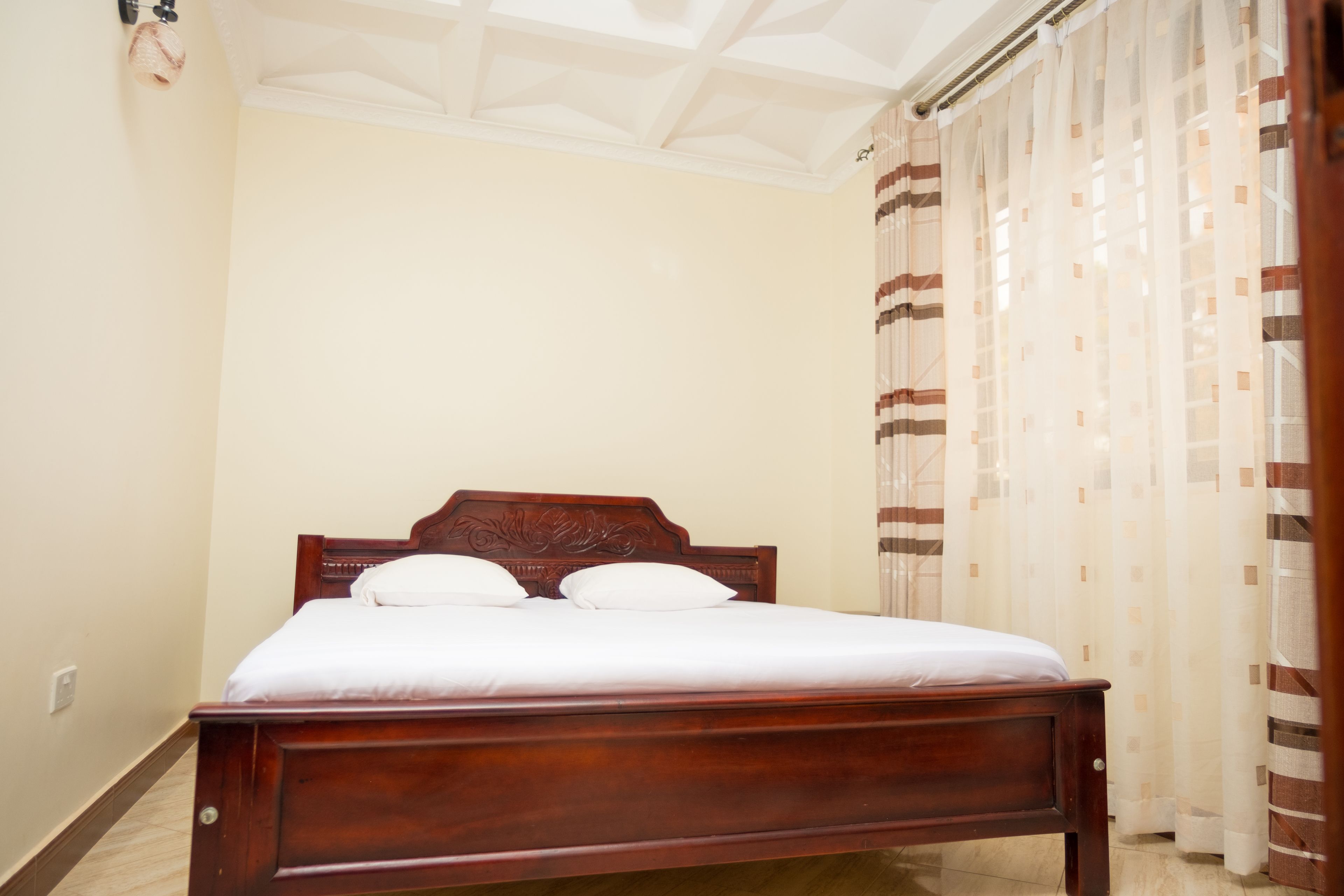 A neatly made bed with a wooden headboard in a room with cream-colored walls and a patterned curtain.