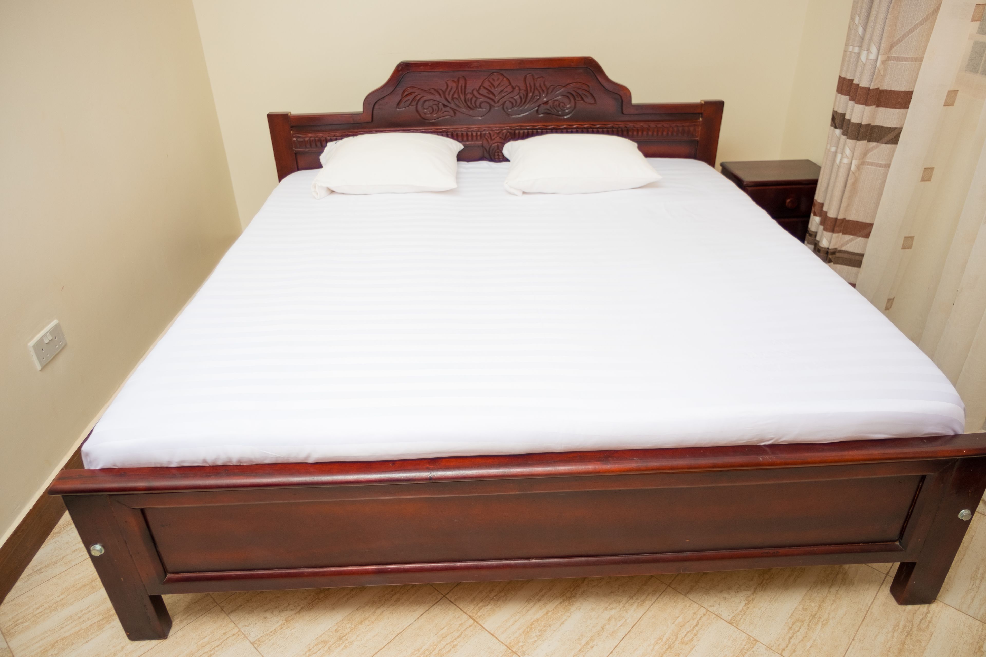 A neatly made bed with a wooden headboard and two pillows, a wooden nightstand beside it, and a curtain partially drawn to the side.