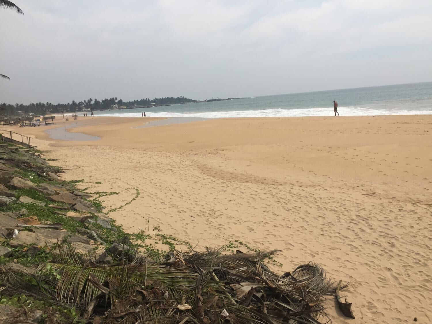 Una playa arenosa con algunas personas caminando, palmeras y un mar tranquilo.
