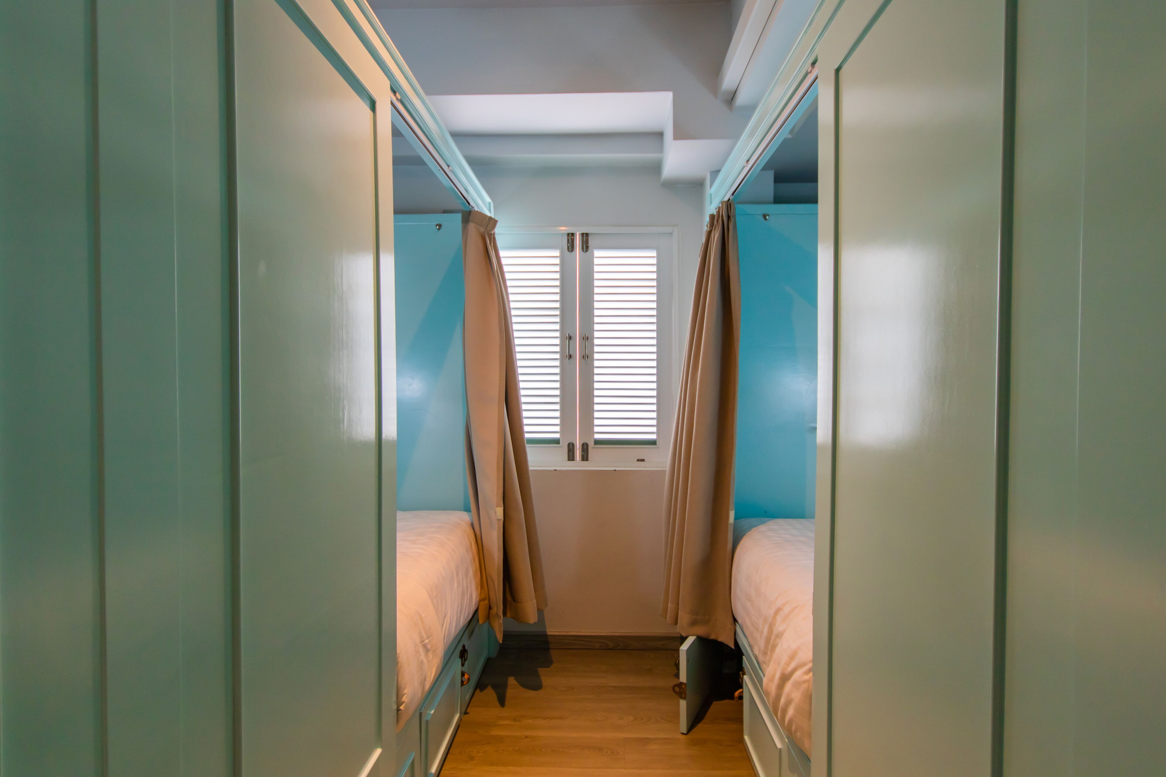A narrow corridor with bunk beds on either side, leading to a window with blinds.