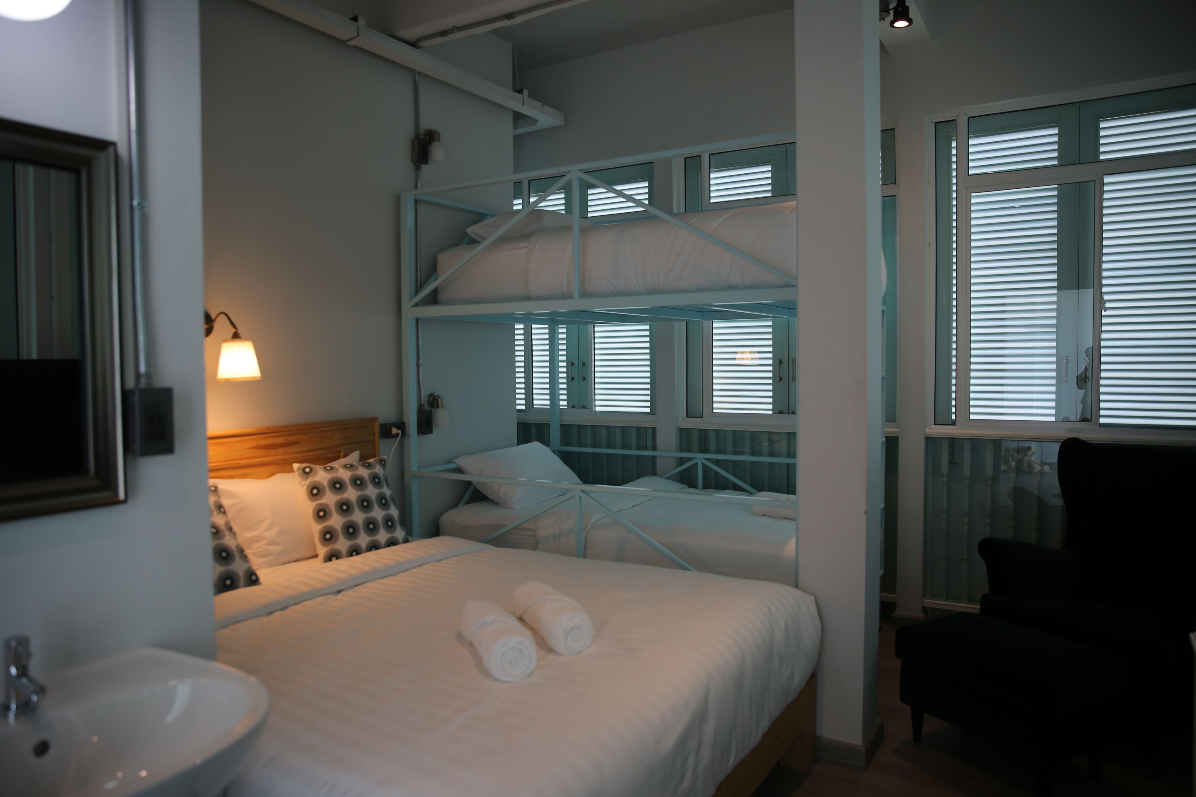 A hotel room with a bunk bed, a bedside table, a lamp, a chair, and a window with blinds.