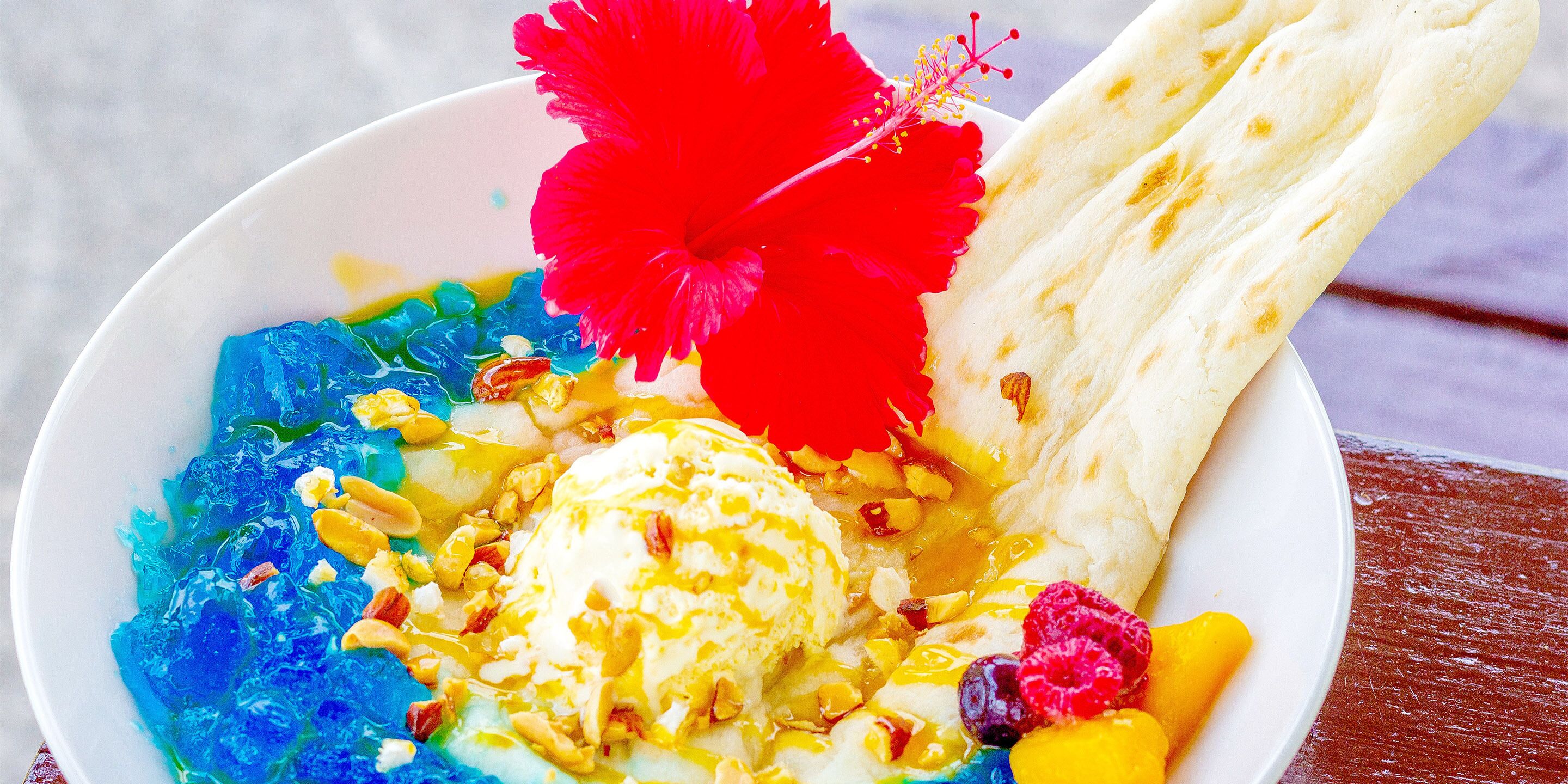 A bowl of dessert with ice cream, fruits, and a red flower.