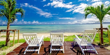 Three white beach chairs on a wooden deck overlooking a beach with clear turquoise water and palm trees.